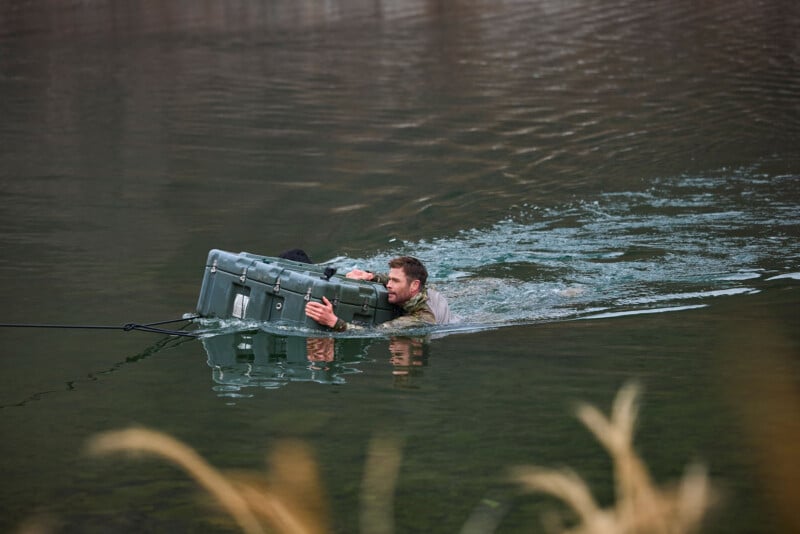 A man swims across a body of water, holding onto a large green plastic container with both arms. He is fully clothed and pulls the container with a rope, with tall grass visible in the foreground.