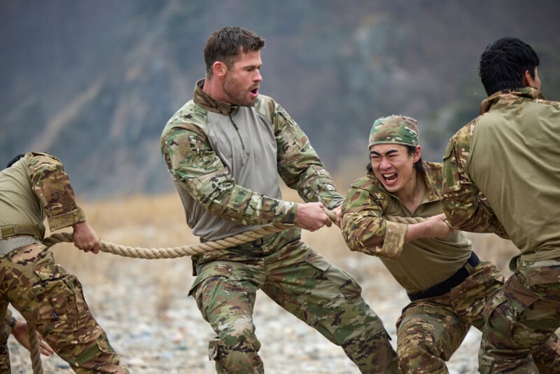 A group of people in camouflage uniforms play tug-of-war outdoors on rocky ground. One person in the middle shouts, while another beside them, wearing a headband, pulls the rope and smiles intensely.