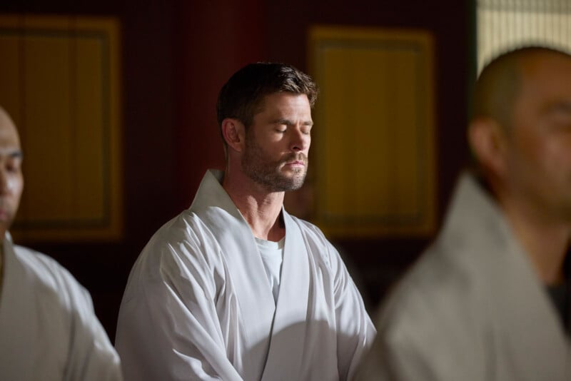 A man in a white robe sits with his eyes closed, appearing to meditate. He is surrounded by others in similar robes, in a softly lit room with blurred background panels.