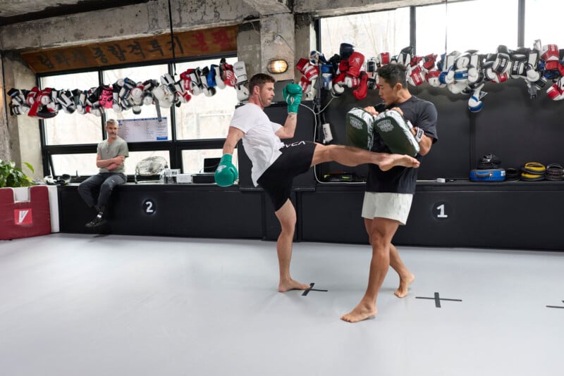 Two men spar in a gym; one delivers a kick to the other's pads. Both wear gloves, with boxing gear hanging on the wall behind them. A third person watches from the side, seated on a bench.