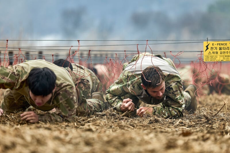 Soldiers in camouflage uniforms crawl on the ground under low wires during a military training exercise. A yellow caution sign with Korean and English text about an electric fence is visible nearby.