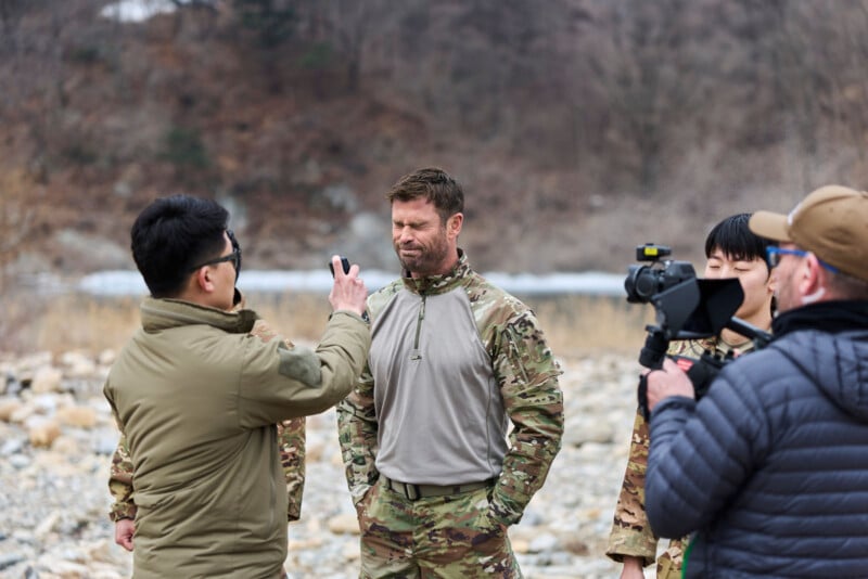 A man in camouflage stands outdoors with eyes closed as another man points a phone at him. Two other people, one with a camera, stand nearby. The scene appears to be a filming or interview set in a rocky, natural area.