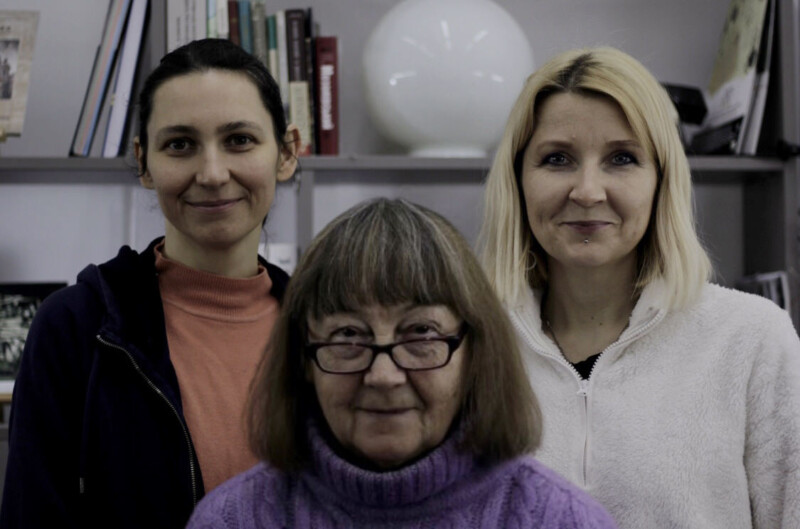 Three women pose indoors in front of shelves with books and a round white lamp. The older woman in front wears glasses and a purple sweater. The two younger women behind her wear dark and light jackets, and all are smiling softly.