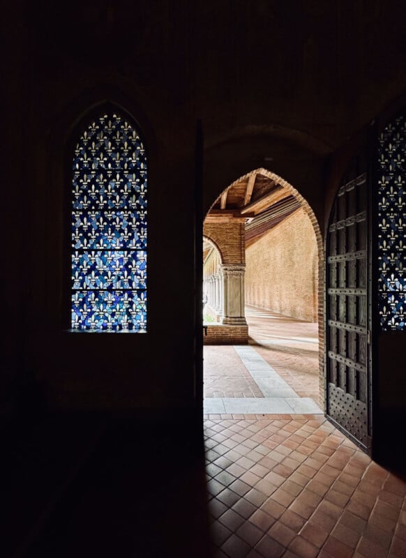 Light streams through an open arched doorway into a stone corridor, next to a tall stained glass window with blue and yellow patterns, creating a contrast between the shadowed interior and sunlit exterior.