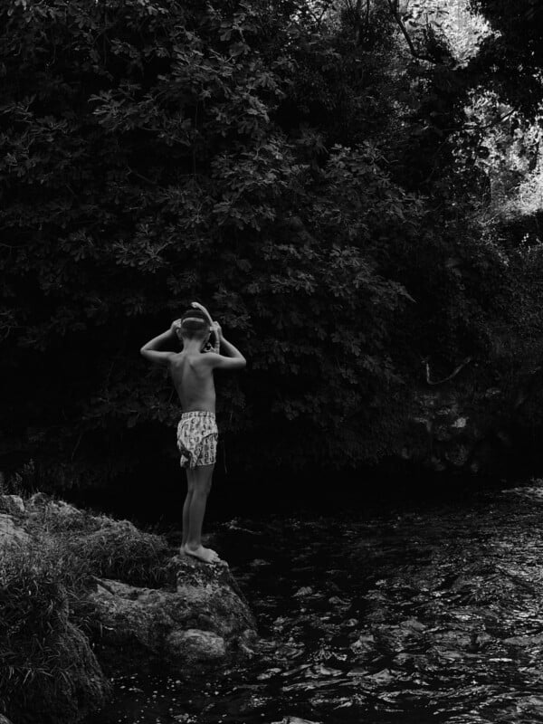A boy in patterned swim trunks stands on a rock by a river, holding his hands to his head, with dense foliage and trees in the background. The scene is in black and white.