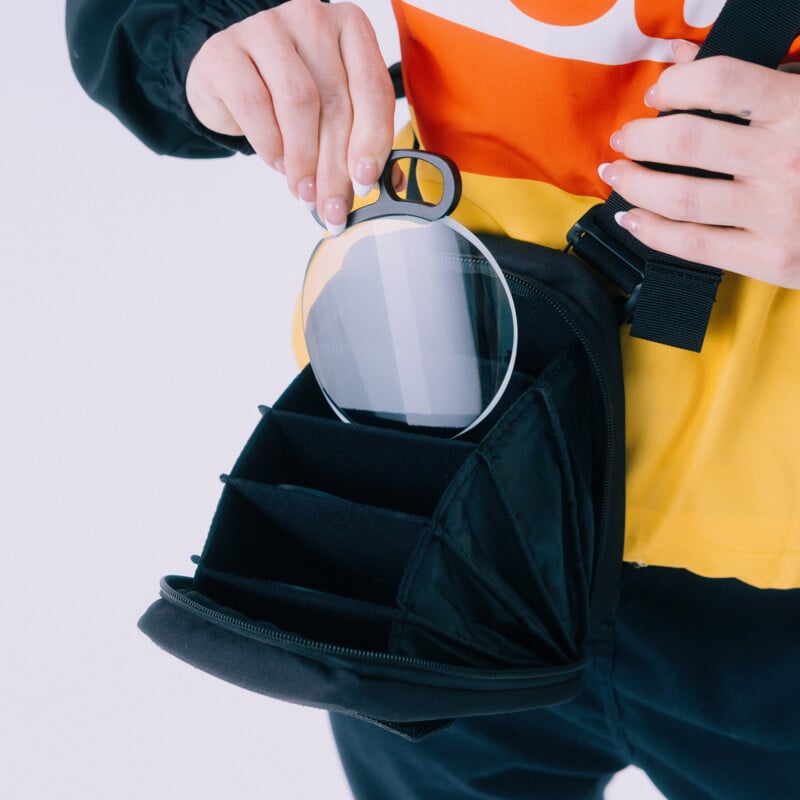 A person wearing a yellow and red top places a large camera lens filter into a black, multi-compartment camera bag.