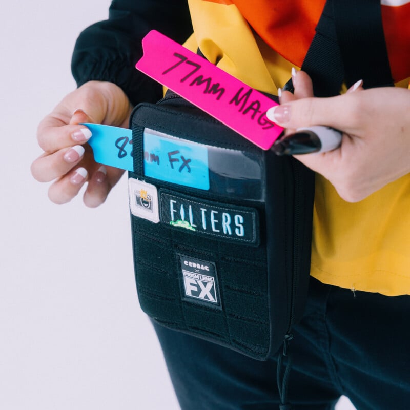 A person holds a pink tag labeled "77mm MAG" and inserts a blue tag labeled "82mm FX" into a black filter case with various filter labels. The person wears a yellow, orange, and black jacket.
