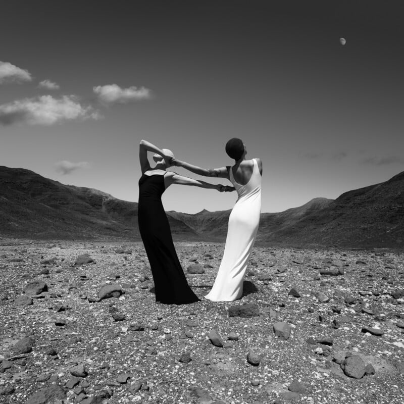 Two women in long, elegant black and white dresses hold hands and lean away from each other on a rocky, barren landscape under a clear sky with the moon visible in the distance.