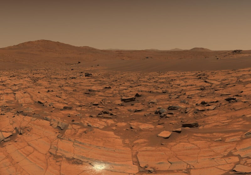 A rocky, reddish landscape on Mars with scattered stones, cracked ground, and distant hills under a hazy, brownish sky. The terrain appears dry and barren.