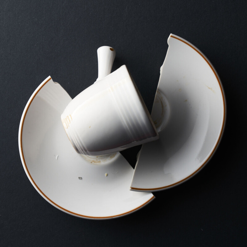 A white teacup sits atop a matching saucer that is broken into two large pieces, all set against a dark background. The saucer has a thin brown rim.