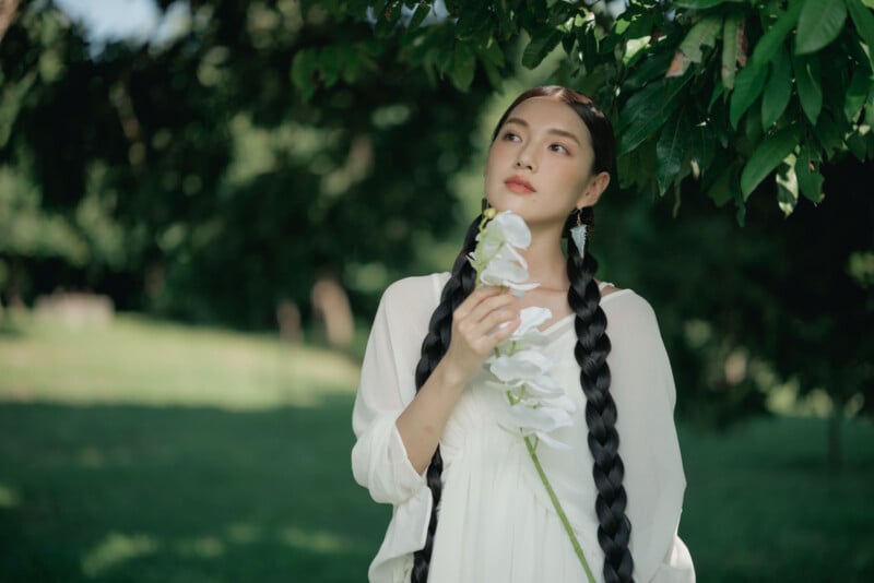 A woman with long braided hair, dressed in a white outfit, holds white flowers and gazes thoughtfully upward while standing outdoors under leafy green branches.