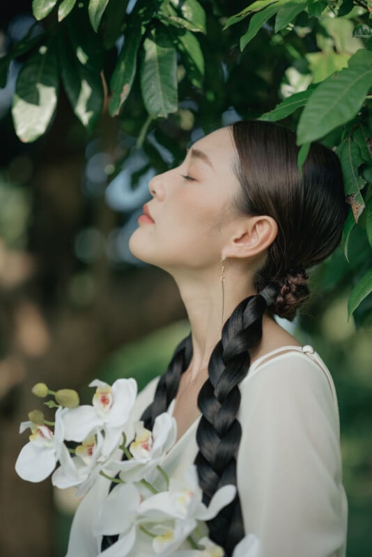 A woman with long, braided hair stands under green leaves, eyes closed, holding white orchids, wearing a white top and gold earring, looking serene in natural light.