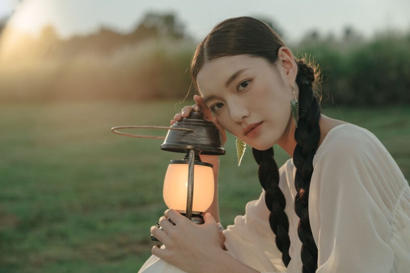A woman with long braided hair, wearing a white top, sits on grass at sunset, holding a lit lantern and looking softly at the camera. The background is blurred greenery.