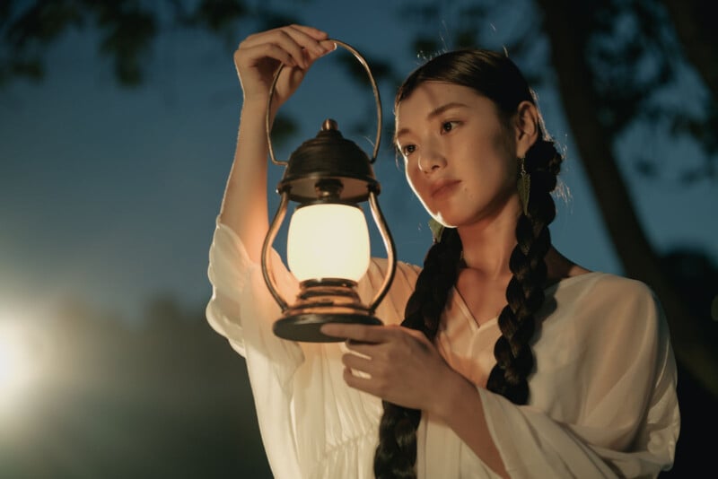 A young woman with long braided hair holds a glowing lantern in the evening, dressed in a white blouse, with trees and a dark sky in the background.
