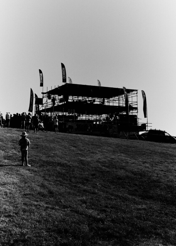 A black and white photo of a grassy hill with a child in the foreground and a large scaffolding structure with flags and crowds gathered near the top under a clear sky.