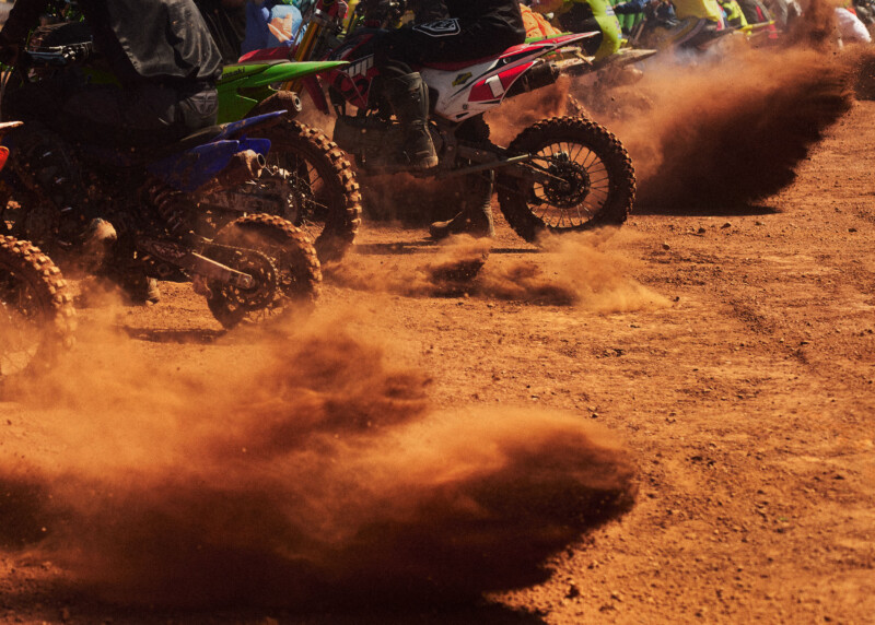 Dirt bike racers on a reddish-brown track kick up clouds of dust as they speed forward, with several motorcycles visible in close proximity and blurred motion suggesting a fast-paced race.