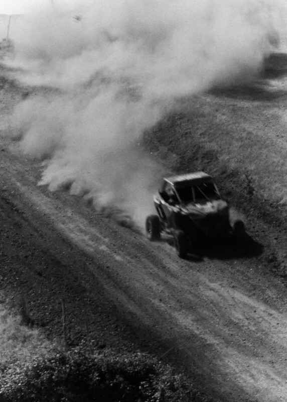 A race car speeds along a dirt track, kicking up a large cloud of dust behind it. The image is in black and white, emphasizing the movement and rugged terrain.