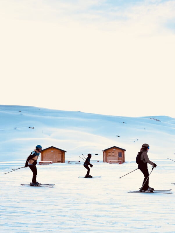 Three people skiing on a snowy slope near two small wooden cabins, with a bright sky and rolling snow-covered hills in the background.