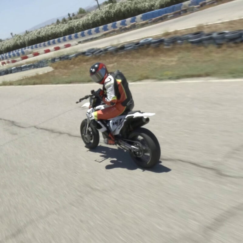 A person in a red and white racing suit and helmet rides a sport motorcycle on a paved race track, leaning into a turn. Tires and barriers line the track in the background.
