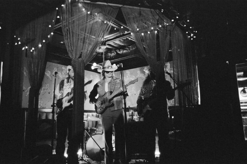 A black-and-white photo of a three-member band performing on stage under string lights. The musicians, including one in a cowboy hat, play guitars in a dimly lit, intimate setting with patterned wallpaper.