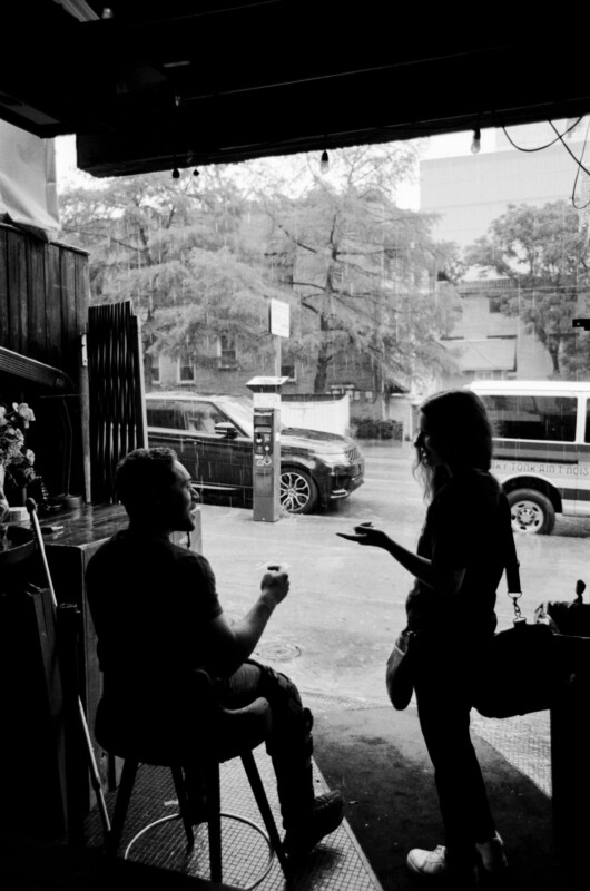 Two people talk in the entrance of a building on a rainy day. One sits on a stool, holding something, while the other stands in front of them, gesturing. Vehicles and trees are visible through the window.