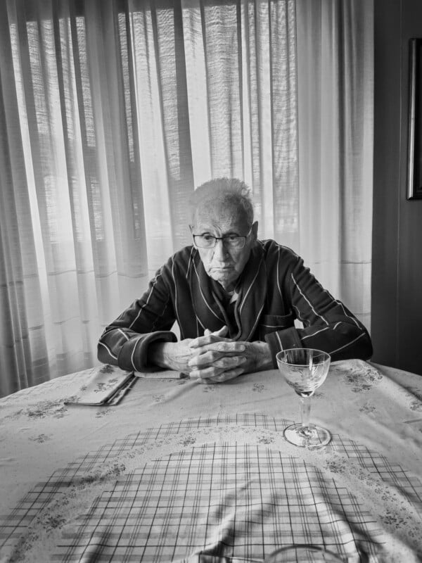 An elderly man in striped pajamas sits alone at a table with his hands folded, looking down. There's a wine glass and folded napkin in front of him. Sheer curtains let in soft light behind him. The photo is in black and white.