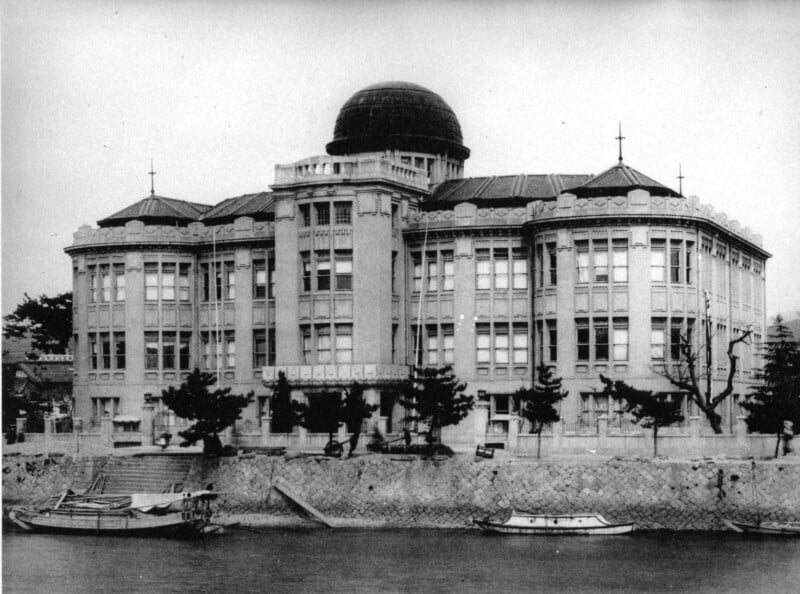 Black and white photo of a large, domed building with many windows, viewed from across a river. Small boats are docked along the stone embankment, and leafless trees line the front of the building.