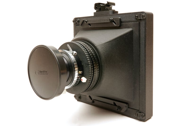 A black large format camera lens assembly with a lens cap, mounted on a square lens board, photographed against a white background.
