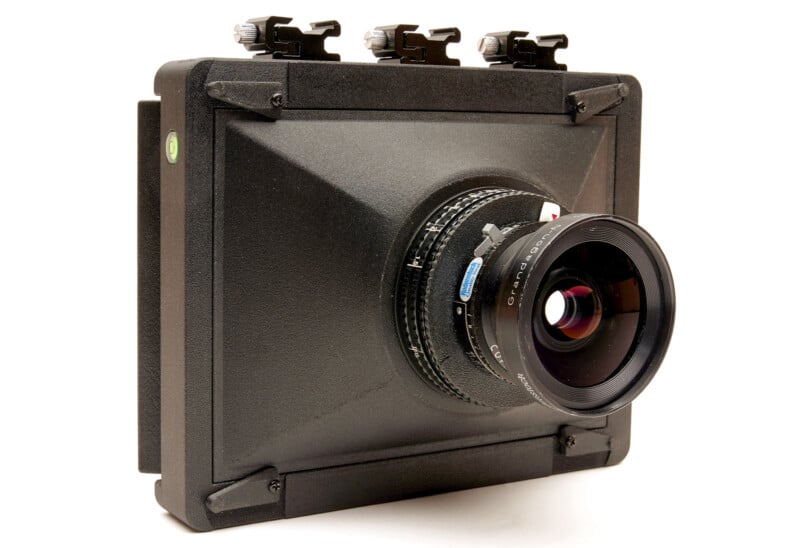 A large format camera with a wide black square body, prominent front lens, and adjustment knobs on top, photographed against a white background.