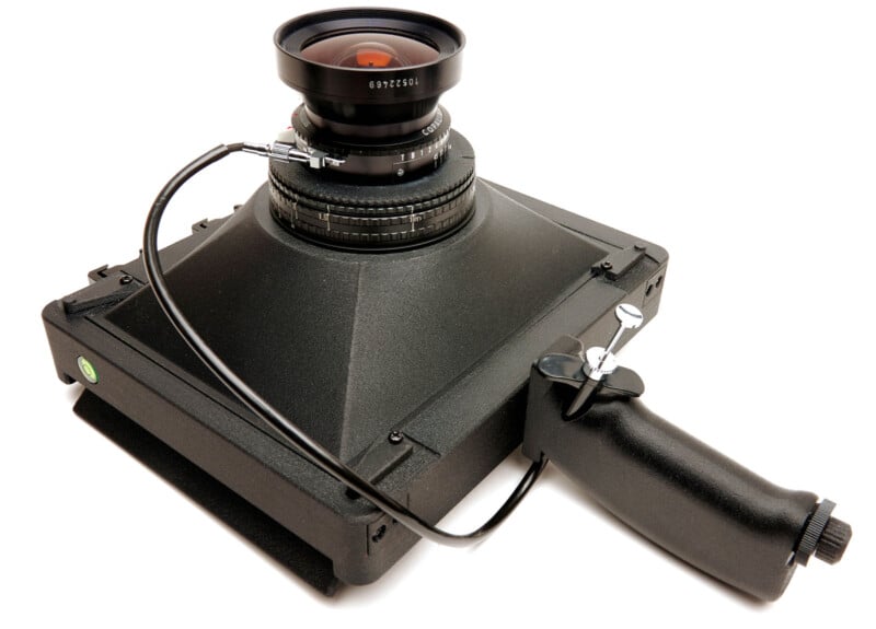 A large format handheld camera with a wide-angle lens, a black handle grip, and a cable release attached to the shutter. The camera body is boxy and matte black.