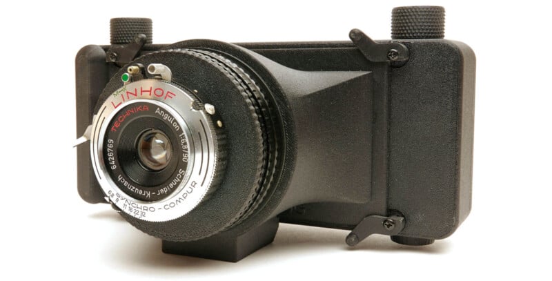 A close-up of a Linhof camera with a Technika Angulon lens, featuring a black, boxy body and metallic details, positioned on a white background.
