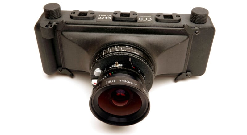 A close-up view of a black camera with a large lens labeled "f=90mm 1:8," showing detailed focus and aperture rings, positioned against a white background.