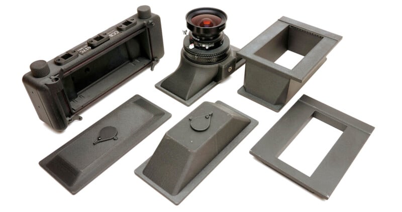 A disassembled large format film camera kit is shown, including the camera back, lens with attached bellows, two rectangular box-like components, and two flat covers, all laid out on a white surface.