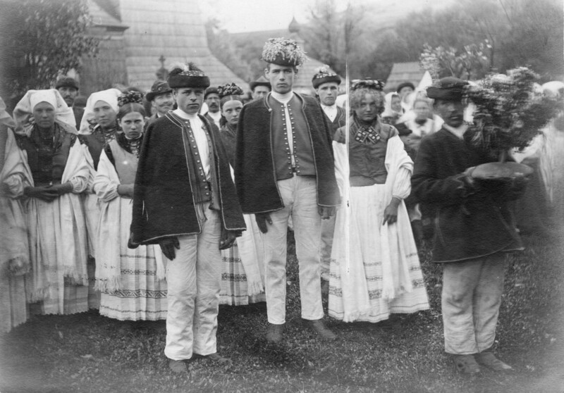 A black and white photo of men and women in traditional folk costumes, standing outdoors in a group. The men wear decorated hats and vests, while the women wear long dresses and head coverings. One person holds a bouquet.