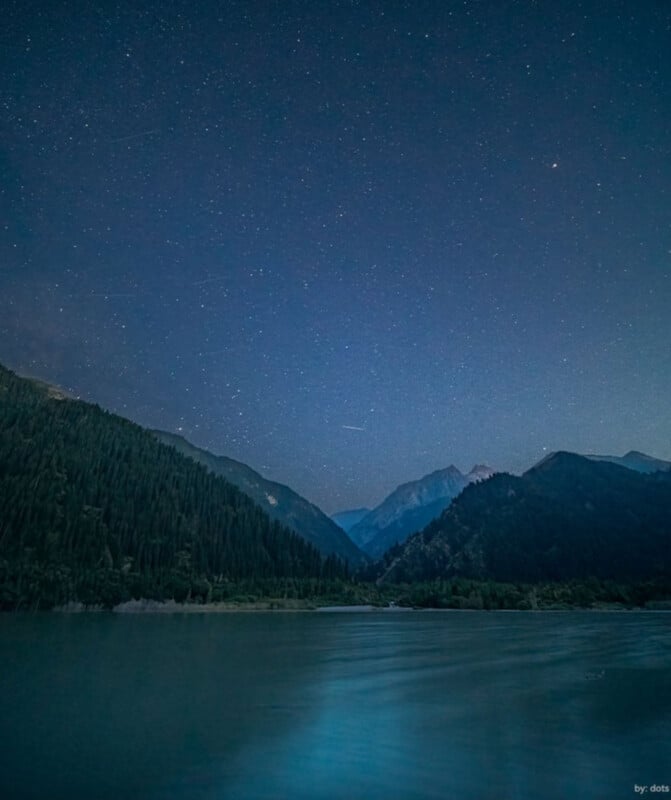 A serene night scene of a lake surrounded by mountains and dense forest, under a clear sky filled with stars. The water reflects the blue tones of the night, creating a peaceful and tranquil atmosphere.