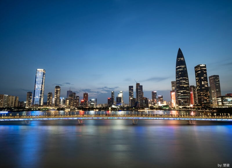 City skyline at dusk with tall modern skyscrapers, colorful lights reflecting on a wide river, and a clear blue sky. A lit pedestrian bridge spans the foreground, adding vibrant hues to the scene.