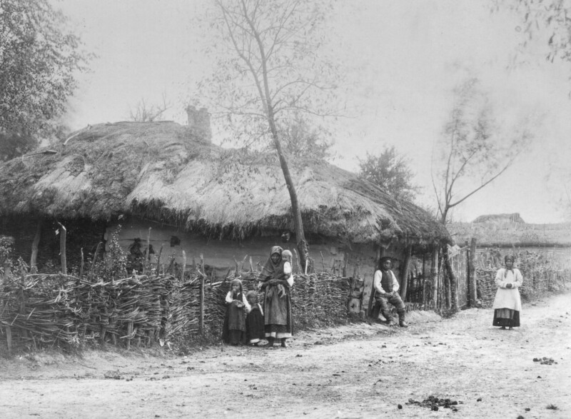 Black-and-white photo of a rural scene with a thatched-roof cottage, a woven fence, and five people—three women, one man, and a girl—standing in front of the house along a dirt road.