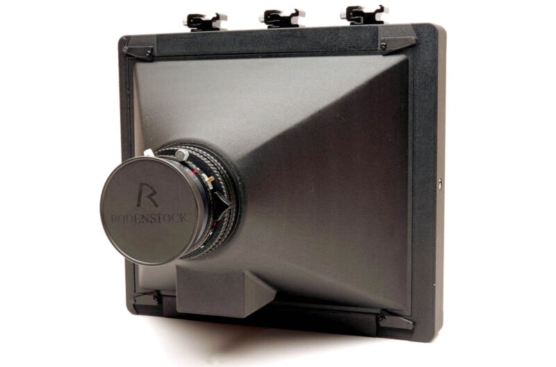 A large format camera lens with a square, black cone-shaped bellows and a Rodenstock lens cap, viewed from an angle against a white background.
