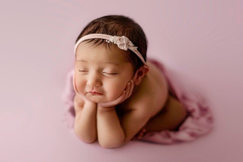 A sleeping newborn baby with closed eyes rests their chin on their hands, lying on a soft pink blanket. The baby wears a pale pink headband with a small flower decoration.
