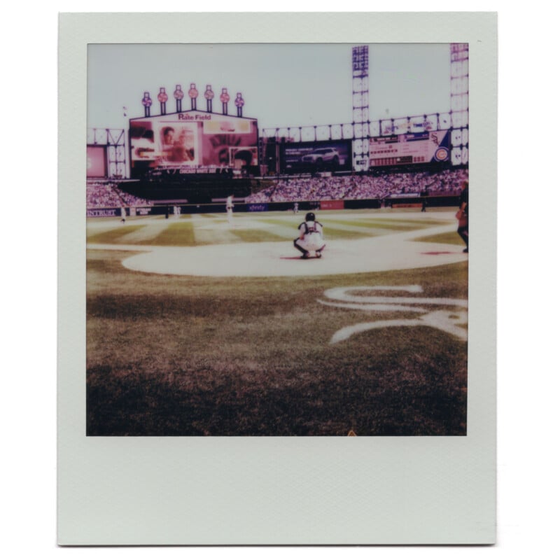 A Polaroid photo of a baseball game at a stadium, showing a batter at home plate, a catcher crouched behind, and a large scoreboard in the background. The stands are filled with spectators.