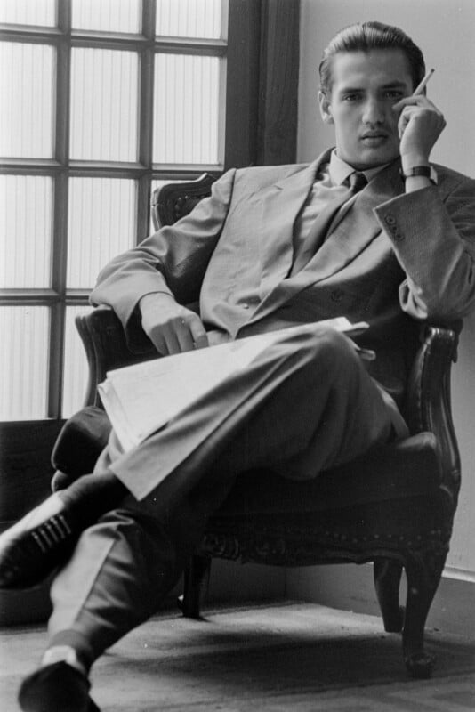 A man in a suit sits in an armchair, legs crossed, holding a cigarette near his face. He gazes thoughtfully into the distance, with a newspaper or document resting on his lap.