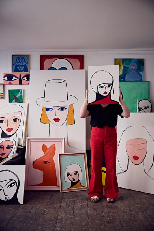 A person in red pants stands in an art studio, holding a painting of a woman's face in front of their head. Colorful, stylized portraits and animal paintings are displayed and stacked around them.
