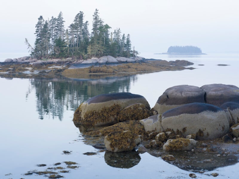 A small rocky island with evergreen trees is reflected in calm water, while large moss-covered rocks sit in the foreground. Another island appears in the misty distance under a pale sky.