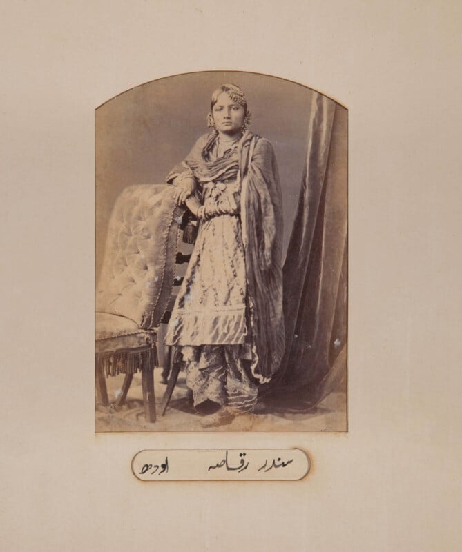 A sepia-toned portrait of a woman in traditional South Asian clothing, standing beside an upholstered chair with one hand resting on it. She looks directly at the camera. Urdu text is written below the photograph.