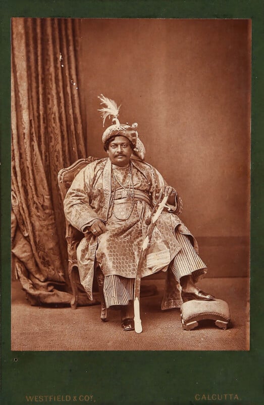A regal man in ornate traditional attire and elaborate jewelry sits on a carved chair, holding a sword. He wears a feathered turban and rests one foot on a cushion. A decorative curtain hangs behind him.