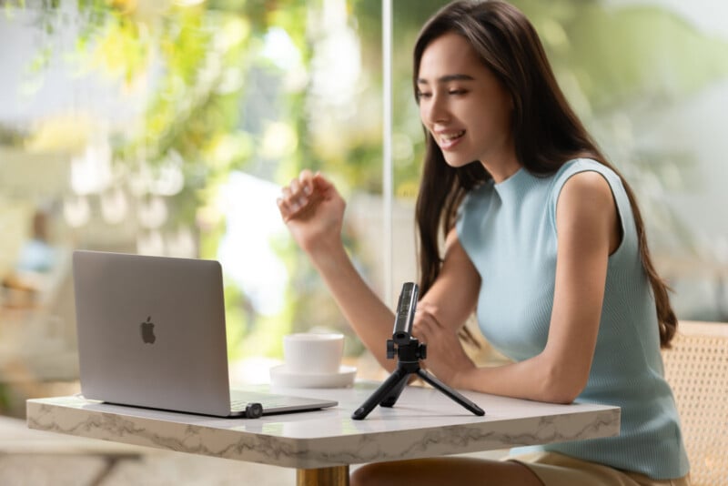A woman sits at a table in a bright cafe, smiling while speaking into a microphone. A laptop, coffee cup, and notebook are in front of her, suggesting she is recording or streaming content.