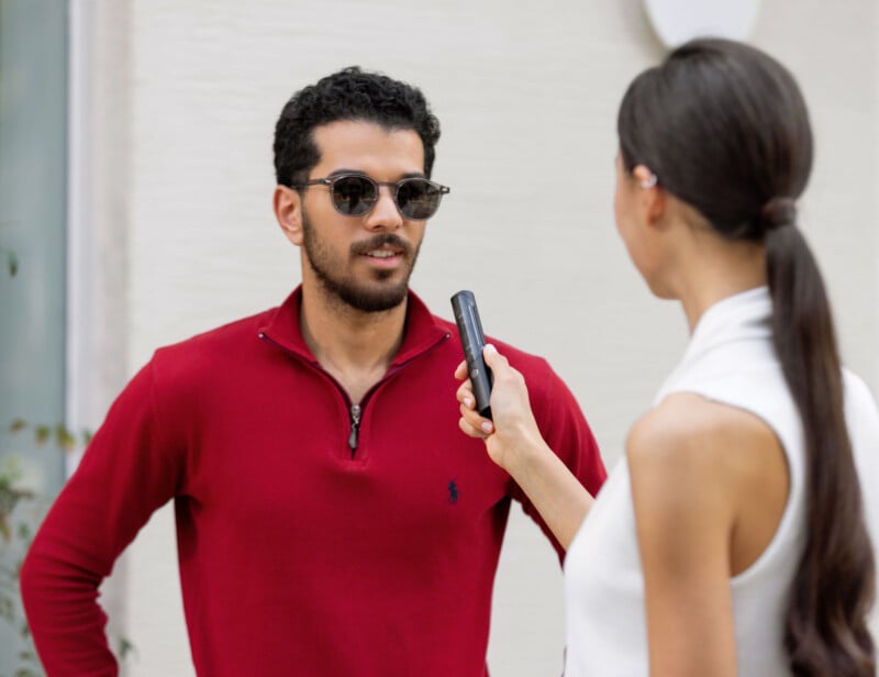 A man in a red long-sleeve shirt and sunglasses is being interviewed by a woman holding a voice recorder. They are standing in front of a light-colored wall.