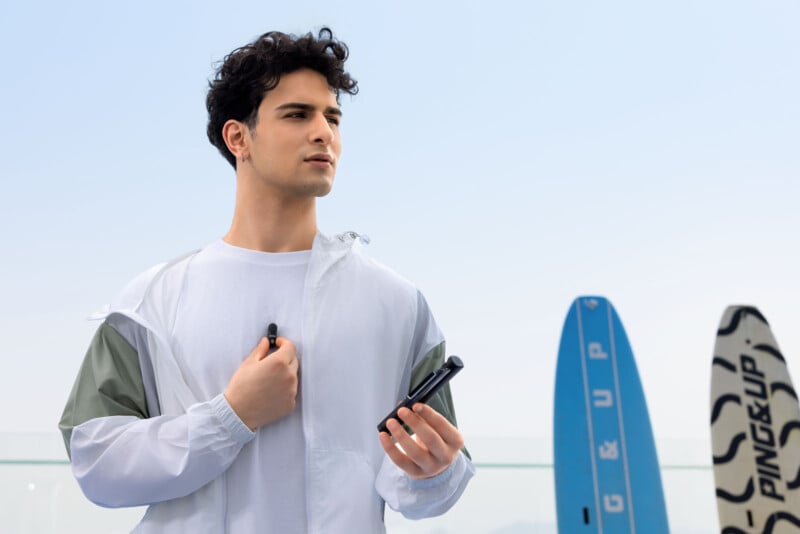 A young man in a white jacket stands outdoors holding a vape device, with two surfboards upright in the background against a clear sky.