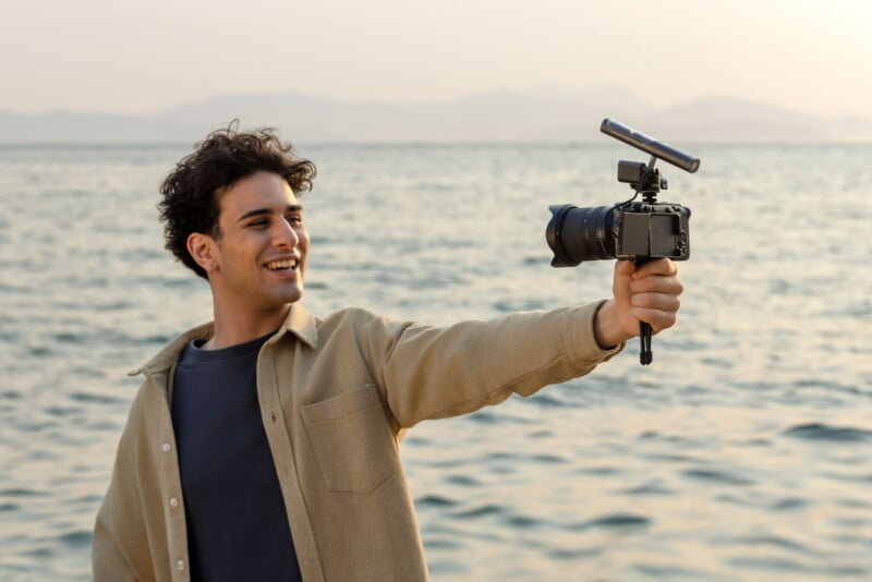 A young man smiles while holding a camera with a microphone, vlogging by the sea during sunset. The background features calm water and distant mountains under a clear sky.
