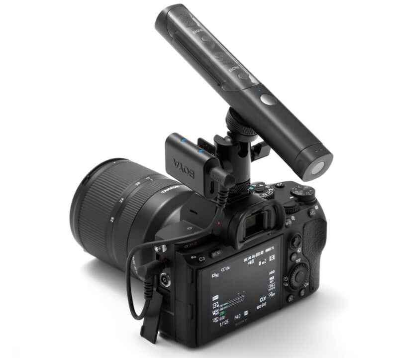 A digital camera with a large lens is shown with an external BOYA microphone attached to its hot shoe mount, aimed forward. The camera's rear LCD screen and control buttons are visible.
